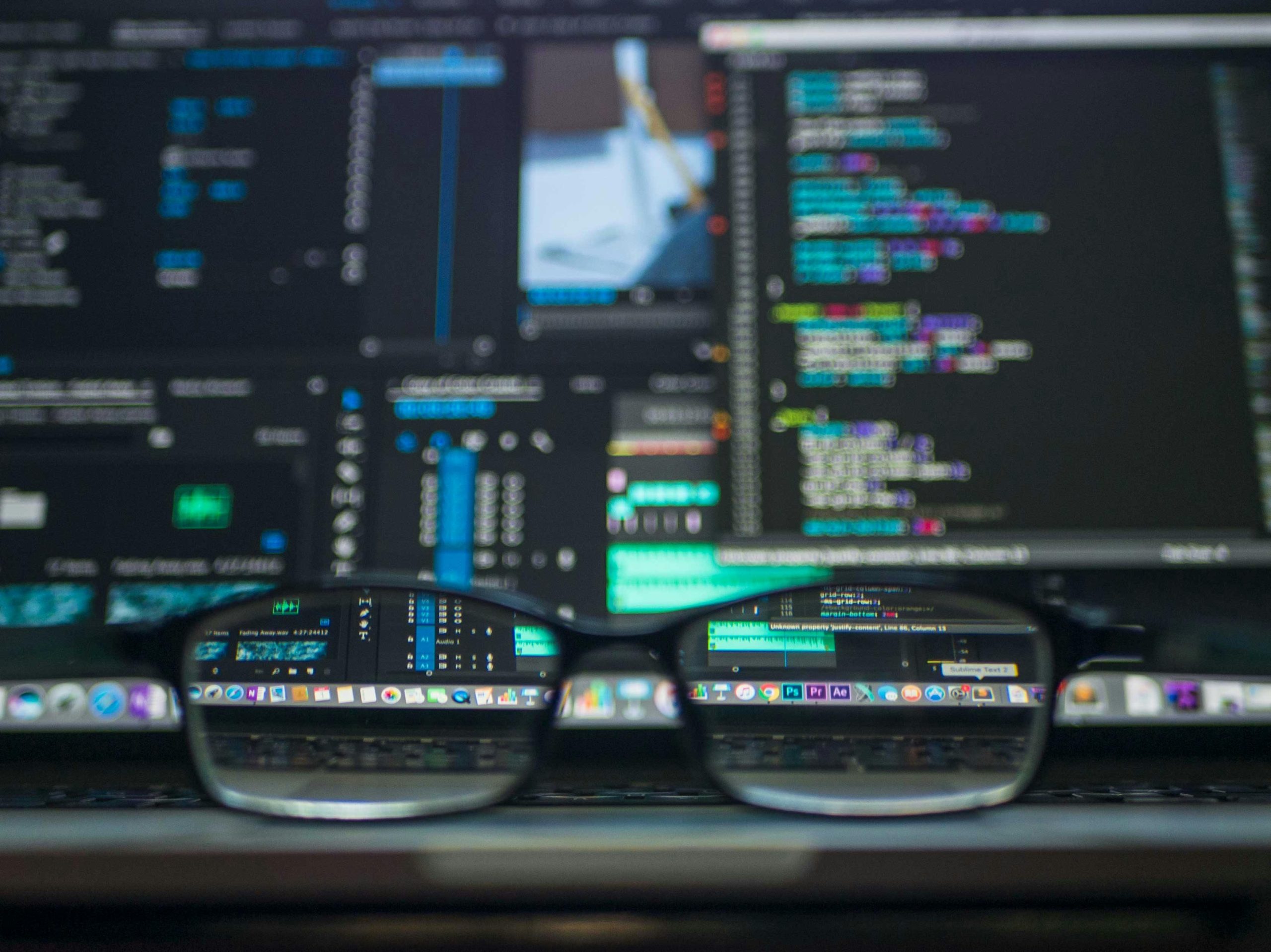 Close-up of eyeglasses placed in front of a laptop screen displaying multiple lines of code and technical interfaces. The code is reflected in the eyeglass lenses.